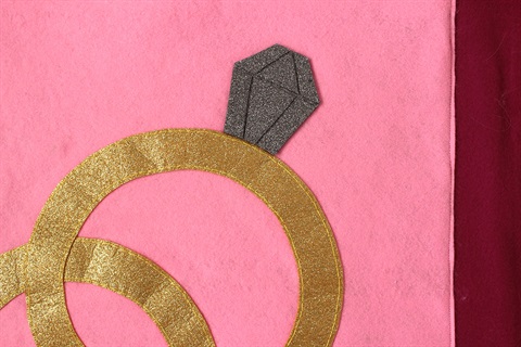 Detail of a felt artwork with a bright pink and burgundy background and two golden ring shapes with a dark grey diamond shape attached